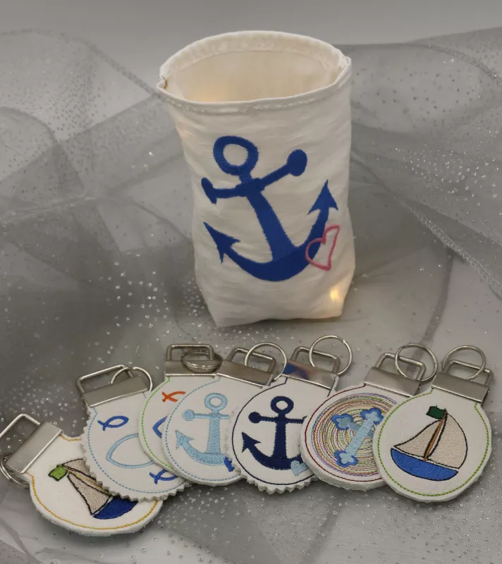 Stickdatei Set christliche Deko und Gastgeschenke (ITH), Foto zeigt hellen Lichtbeutel mit Ankeremblem, davor liegen 7 runde Anhänger. Diese mit angeklammerter Metallöse mit Schlüsselring und diversen Symbolen (Segelboot, Fische, Anker, Kreuz).