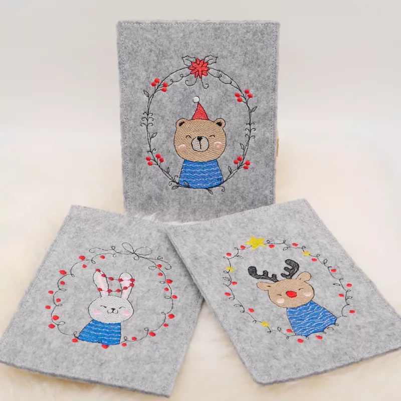 Stickdatei Set ITH Christmas-Cards - Grußkartenhüllen, Foto zeigt 3 Hochkant-Karten aus hellgrauem Filz. Bunte Bestickung mit jeweils diversem Blütenkranz, darin unterschiedliche Motive (Teddybär, Häschen, Elch).