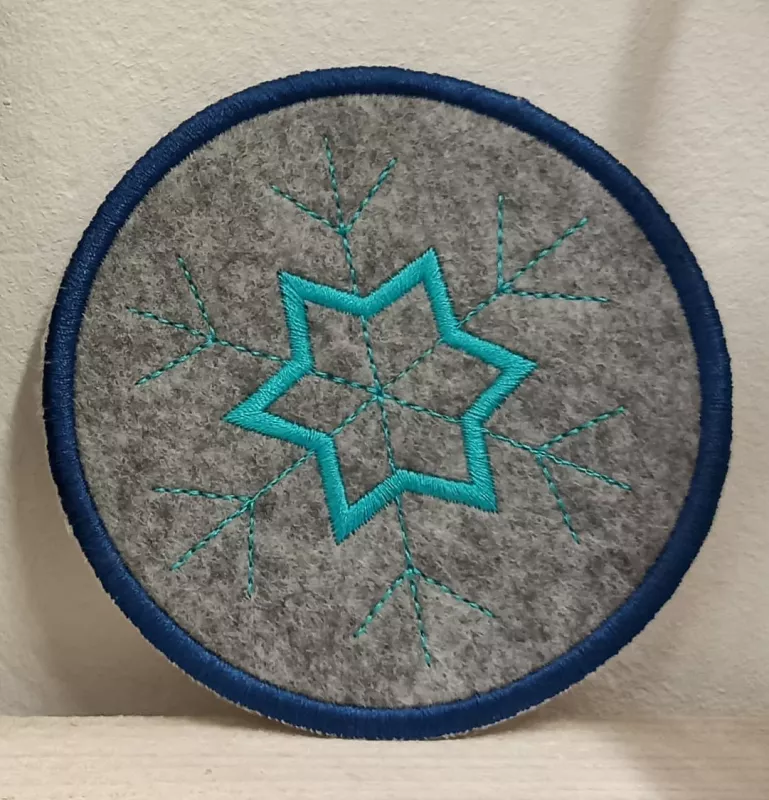 Freebie Stickdatei ITH Mug Rug Frohe Weihnachten (10x10 und 13x18), kreisrund aus grau-meliertem Filz mit dunkelblauer Umrandung. Innen Motiv Eiskristall in türkis. 