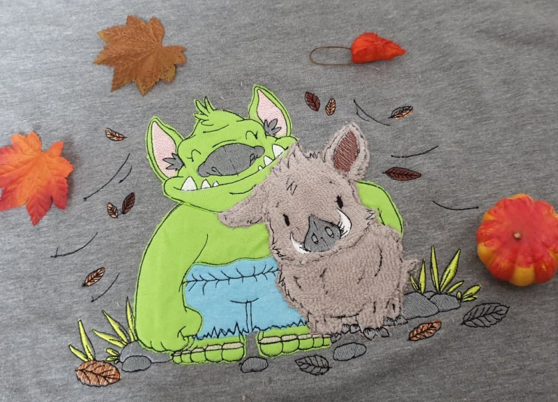 Stickdatei Trollfreundschaft Applikation (5 Größen), Foto mit Motiv auf graubraunem Stoff. Grüner Troll mit hellblauer Hose umarmt hellbraunes Wildschwein. Umgeben von gestickten Blättern, Steinen, Grasbüscheln. Stickdatei Trollfreundschaft Applikation (5 Größen), Foto mit Motiv auf graubraunem Stoff. Grüner Troll mit hellblauer Hose umarmt hellbraunes Wildschwein. Umgeben von gestickten Blättern, Steinen, Grasbüscheln.