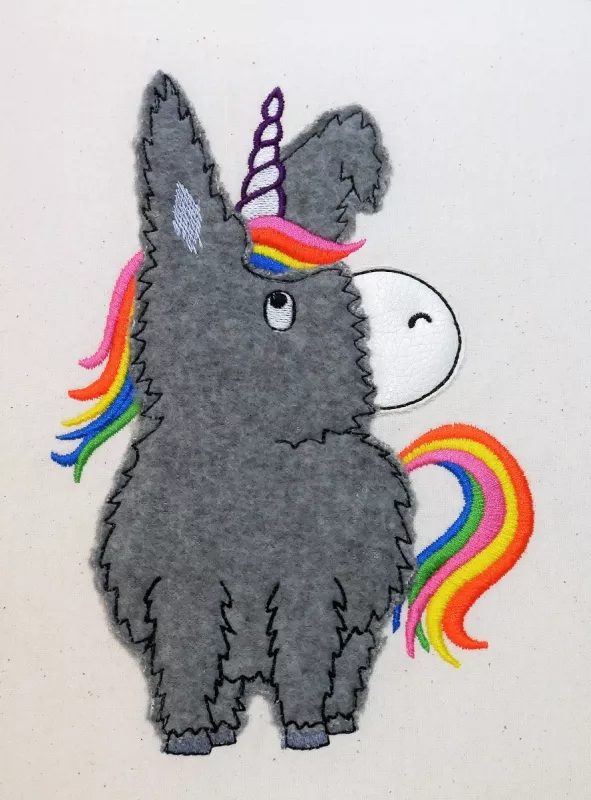 Stickdatei Donkeycorn Doodle Applikation in 5 Größen, Foto zeigt Motiv auf hellem Stoff. Esel aus dunkelgrauem Flauschstoff, weiße Schnauze, mit hellem Horn. Mähne und Schweif in bunten Farben gestickt.