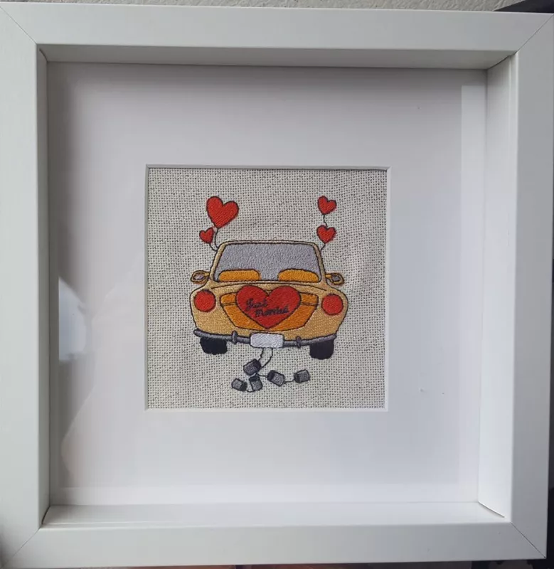 Stickdatei Set Hochzeit, Foto zeigt Motiv in Bilderrahmen. Oldie-Auto, Herzluftballons daran angebunden und Konservendosenkette. Trägt Herz mit Schriftzug "Just married".