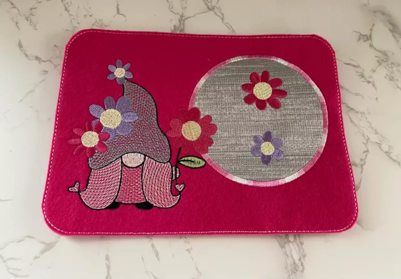 Stickdatei Set Gnom Mug Rugs 4 Jahreszeiten + Einzelmotive, Foto mit rechteckigem Tassenteppich aus rotem Filz im Querformat auf Tisch platziert. Linksseitig Gnom mit Blüten und Blume in der Hand (Farbtöne lila, rosa, gelb, rot). Rechtsseitig kreisrunder