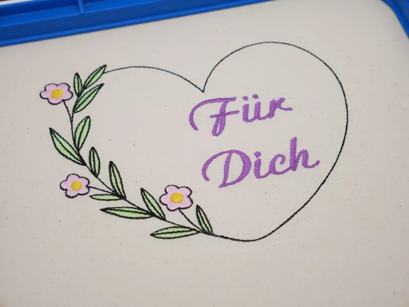 Stickdatei Set florale Rahmen + Schriftzüge, Foto mit in Stickrahmen eingespanntem hellen Stoff. Darauf Herzumriss mit Schriftzug "Für Dich" im Inneren. Herz wird auf einer Seite durch einen Blüten-Blätter-Zweig verstärkt.