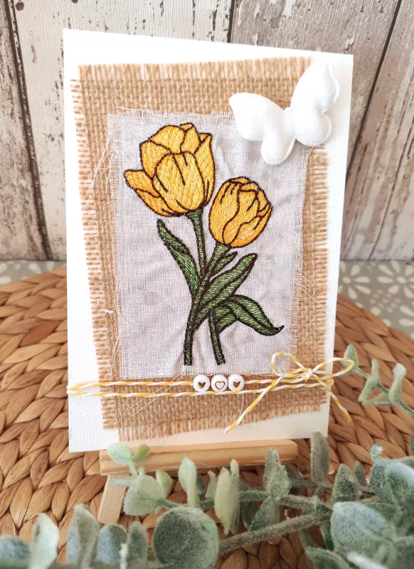 Stickdatei Set 6 Blumen für den 10x10 & 13x18er Rahmen, Foto zeigt Motiv auf weißem Untergrund als hochkantiges Bild, auf Staffelei stehend. Grober juteartiger gewebter Unterstoff mit weißem mullartigem grob gewebtem Stoff darauf. Mit Motiv gelbe Tulpen m