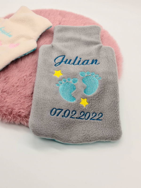 Stickdatei XXL Set ITH Wärmflaschenhüllen mit 24 Designs. Foto zeigt braungraue Hülle mit Auszug für Flaschenhals, Schriftzug "Julian 07.02.2022" in blau, türkise Babyfüße, gelbe Sternchen. Im Hintergrund anteilig weitere Hülle erkennbar.