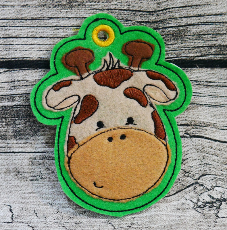 Stickdatei Giraffe Applikation + ITH Anhänger, Foto zeigt mittig oben geösten Giraffenkopfanhänger auf hellem Holzuntergrund. Filzgrund in grün, Bestickung: Brauntöne, Vanille.