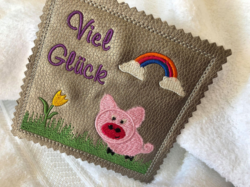 Stickdatei Set ITH Mini-Grußkarten inkl. Abwandlung zur Geschenktasche, Foto mit 1 quadratischen Ausarbeitung, präsentiert auf hellem Untergrund. Braunes Kunstleder mit bunter Bestickung Schweinchen, Regenbogen, Wiese mit Tulpe, Schriftzug "Viel Glück".