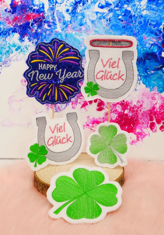 Stickdatei Set Glück & Silvester Stabdesigns , Foto mit 5 Designs, präsentiert auf Holzscheibe, vor buntem Hintergrund.1 mal Lichtschein Silvesterrakete mit Schriftzug "Happy New Year"; 2 mal Glückskleeblatt mit Stiel links bzw. rechts; 2 mal Hufeisen mi