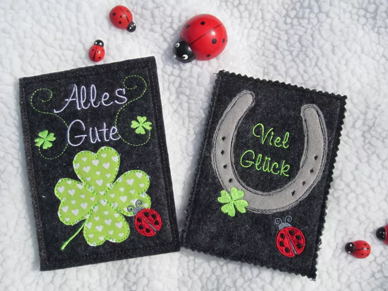 Stickdatei Set ITH Glückwunschkarten inkl. Abwandlung zur Geschenktasche (div. Rahmengr.), Foto von 2 anthrazitfarbenen Hochkant-Karten, auf hellem Untergrund liegend, umgeben von Deko-Marienkäfern. Eine mit Glückskleeblättern und Marienkäfer, Schriftzug