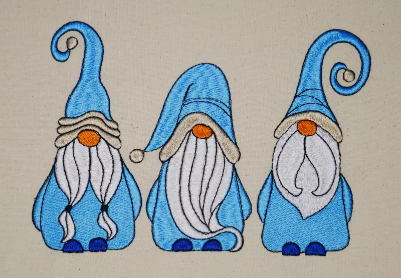 Stickdatei Set Gnomtrio - drei Gnome in Vollstick mit ITH-Lichtbeutel-Dateien (13x18), Foto mit Gnom-Trio auf hellem Stoff. Gnome überwiegend in blau-weiß gehalten.