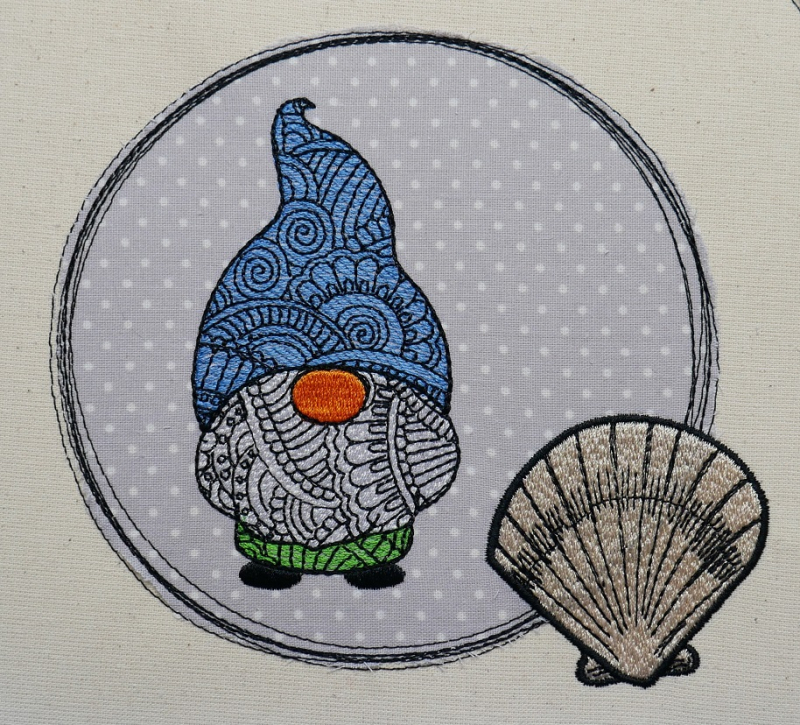 Gnom mit Mandalamuter und blauer Mütze auf hellgrauem Applikationsstoff mit weißen Punkten in einem Kreis verziert mit einer Muschel