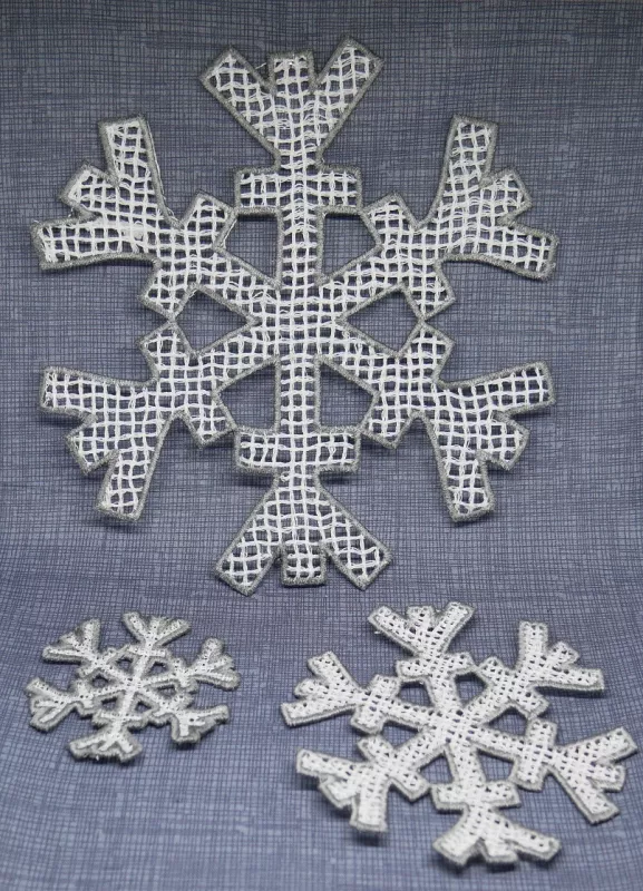 Stickdatei FSL Schneeflocken-Set 6,1 x 6,3 / 9,6 x 9,9 / 17 x 17,5 cm, Foto dreier heller Ausfertigungen, auf grauem Stoff liegend. Diverse Größen, Strukturen differieren von eher massiv zu gröber werdender Netzartigkeit.