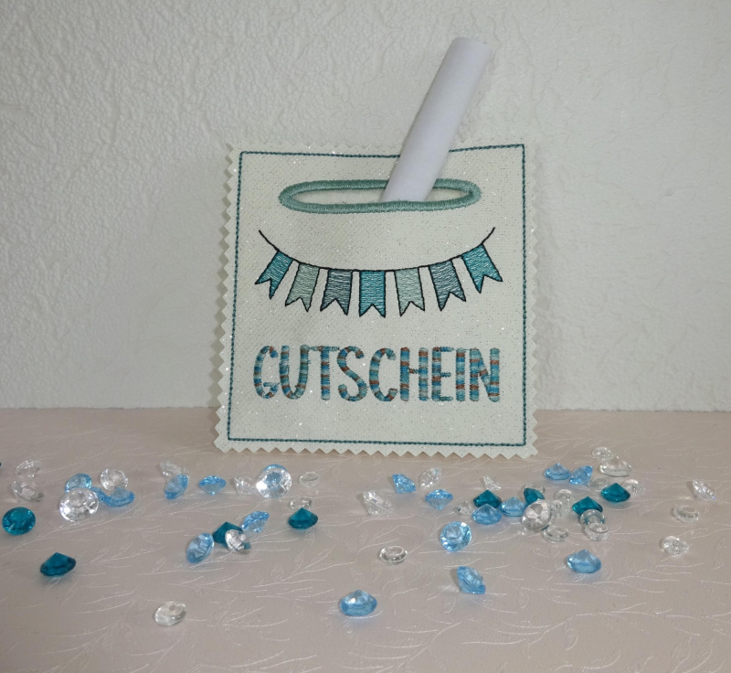 Stickdatei Set ITH Wunscherfüller für alle Gelegenheiten, Foto zeigt auf dekoriertem Untergrund ein ausgefertigtes Exemplar, aus dem ein gerolltes Papier herausragt. Quadratische helle Tasche, in Grün-Blau-Tönen bestickt mit Fähnchengirlande, breite Einst