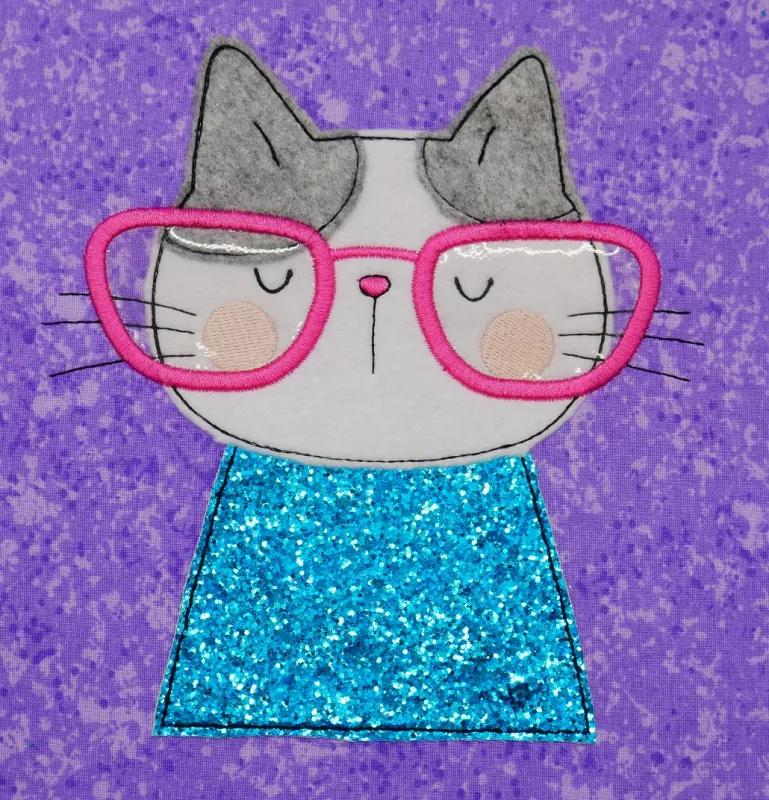 Stickdatei Set I'm Happy Cat Doodle Applikation inkl. ITH Anhänger, Foto zeigt Katzenbüste als Applikation auf lila-gemustertem Stoff. Rosarote Brille, Katzenfarben weiß, grau und glitzerblau.