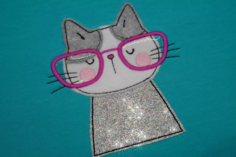 Stickdatei Set I'm Happy Cat Doodle Applikation inkl. ITH Anhänger, Foto zeigt Ausfertigung auf petrolfarbenem Stoff. Katzenportrait aus grauem, weißem und Glitzer-Stoff, mit rosa-roter Brille.