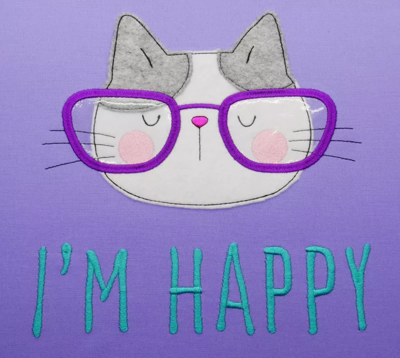 Stickdatei Set I'm Happy Cat Doodle Applikation inkl. ITH Anhänger, Foto zeigt Applikation auf lila Stoff. Katzenkopf aus grauem und weißem Material, fuchsiafarbener Brille. Darunter Schriftzug "I´m happy".