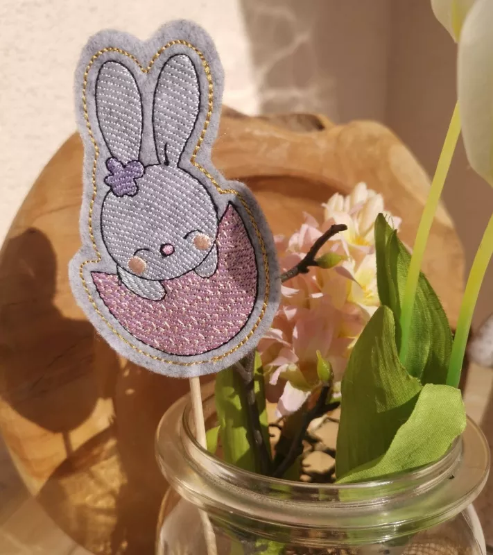 Stickdatei Set Osterhasen ITH Stabdesigns, Foto mit Designstab, der in einem Glas mit Frühlingsblumen steckt. Osterhase auf grauem Filz, anteilig zu sehen, sitzt in einer halben Ostereierschale. Schmückung an einem Ohr.