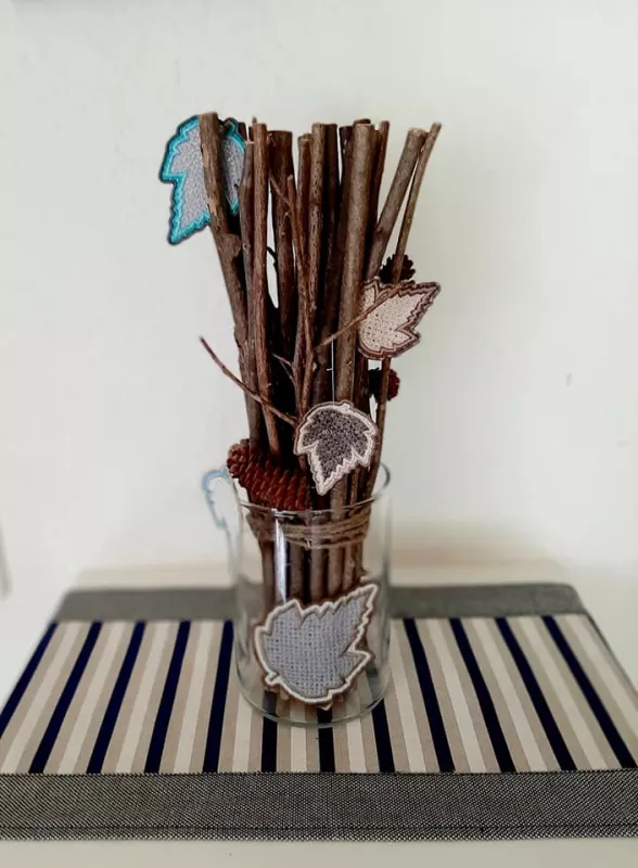 Stickdatei Set FSL Gnome rund um das Jahr + ITH Anhänger, Foto zeigt 4 einzeln ausgestickte Blätter, eingesteckt zwischen Dekoästen in einem Glas. Blätter in netzartiger Optik gefertigt, je zweifarbig (Innenfläche und Umrandung), unterschiedliche Farben.