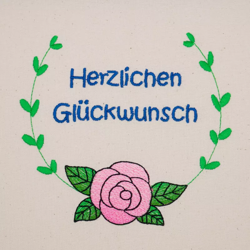 Stickdatei Set florale Rahmen + Schriftzüge,  Foto mit Bestickung in grün-rosa-blau auf hellem Stoff. Von einer Rose mit Blättern führen 2 gebogene Blattzweige randgebend nach oben, dort Rahmen offen. Innen der Schriftzug "Herzlichen Glückwunsch".