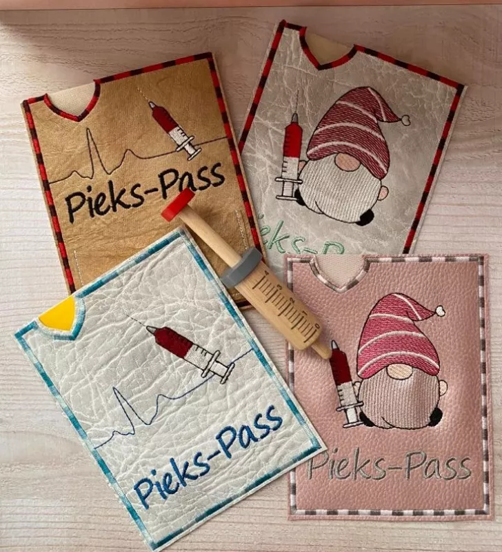 Stickdatei Set ITH Impfpass-Hüllen 24 Designs (13x18), Foto mit 4 beispielhaften Hüllen, auf Holzuntergrund liegend, darauf Holzspritze als Accessoire. 2 mal Spritzengnom; 2 mal EKG-Kurve mit Spritze. Alle zusätzlich mit Schriftzug "Pieks-Pass".