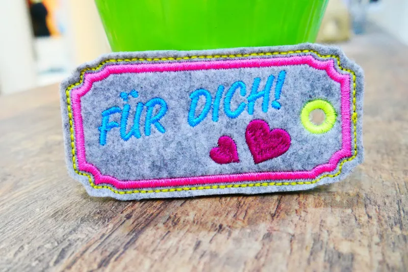 Freebie Stickdatei ITH Anhänger Für Dich (10x10), Foto des ausgestickten Anhängers in länglichem zierrahmenartigem Querformat aus bläulich-grau meliertem Filz. Am Rand gelb abgenäht, darin rosa Stickumrandung, gelbe Garnöse seitlich, 2 rote Vollstick-Herz