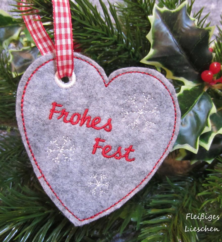 Stickdatei Set ITH Anhänger Frohes Fest, Foto zeigt 1 geösten Anhänger aus grauem Filz am Bändchen hängend, Hintergrund Weihnachtsdeko. Schriftzug "Frohes Fest", Form: Herz.