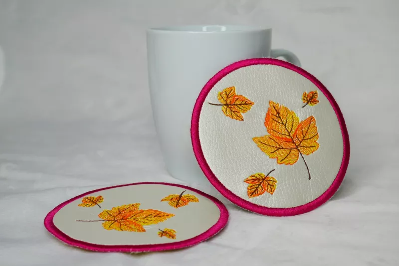 Freebie Stickdatei ITH Mug Rug Herbst (13x18 und 10x10 Rahmen), Foto zweier identischer runder Untersetzer mit Tasse als Deko-Element. Kreisrund aus hellem Kunstleder, rot umrandet, darin 4 Herbstblätter in orange-gelb-dunkelbraun.