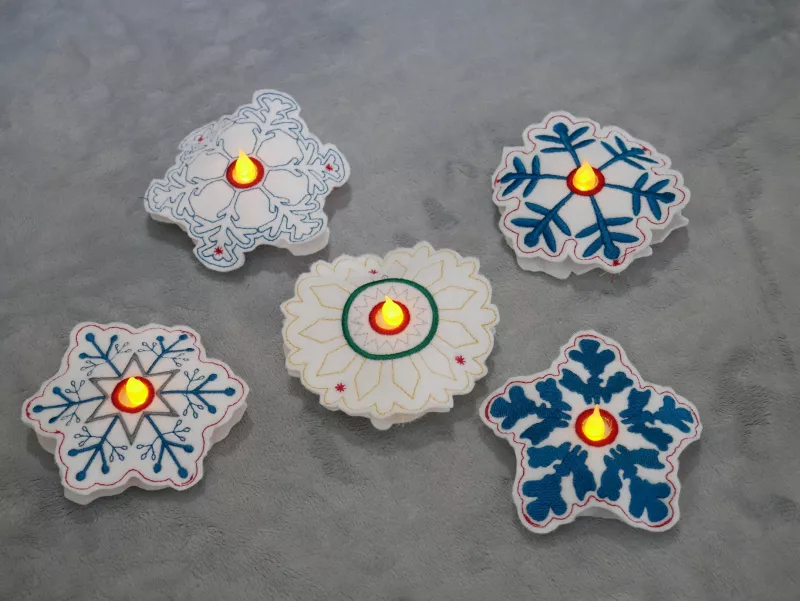 Stickdatei Set ITH Schneeflocken in 2 Größen, Foto zeigt 5 Schneeflocken, präsentiert auf hellem Untergrund. präsentiert. Zentrum LED-Licht-beleuchtet. Stickornamente divers, hauptsächlich blau-weiß.