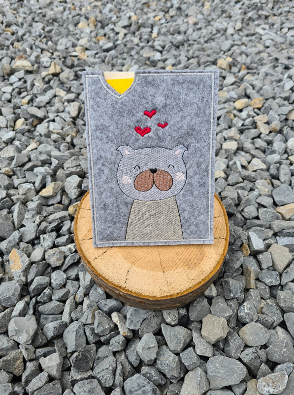 Stickdatei Set ITH TIERPASS-Hüllen 13 Designs (13x18), Foto, das eine Hülle auf einer Holzscheibe präsentiert, die auf grauem Kieselsteinuntergrund steht. Bestickung mit Hund und Herzchen, Hülle aus grauem Filz.