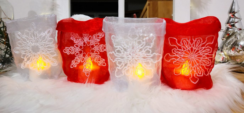 Stickdatei Set ITH Lichtbeutel mit Wintermandalas (13x18 und 16x26), Foto mit 4 nebeneinander auf weißem Fell stehenden leuchtenden Lichtbeuteln, rot und transparent-weiß. Auf jedem befindet sich eine mandalaartige helle Stern-Bestickung, die Oberseiten d