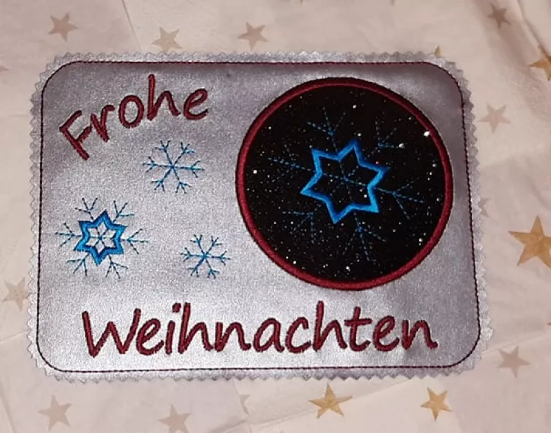 Freebie Stickdatei ITH Mug Rug Frohe Weihnachten (10x10 und 13x18), querformatiger Tassenteppich, silbrig-metallischer Untergrund. Schriftzug "Frohe Weihnachten" und Umrandung des Abstellplatzes in rot, blaue Stickereien Eiskristalle. Abstellplatz kreisru