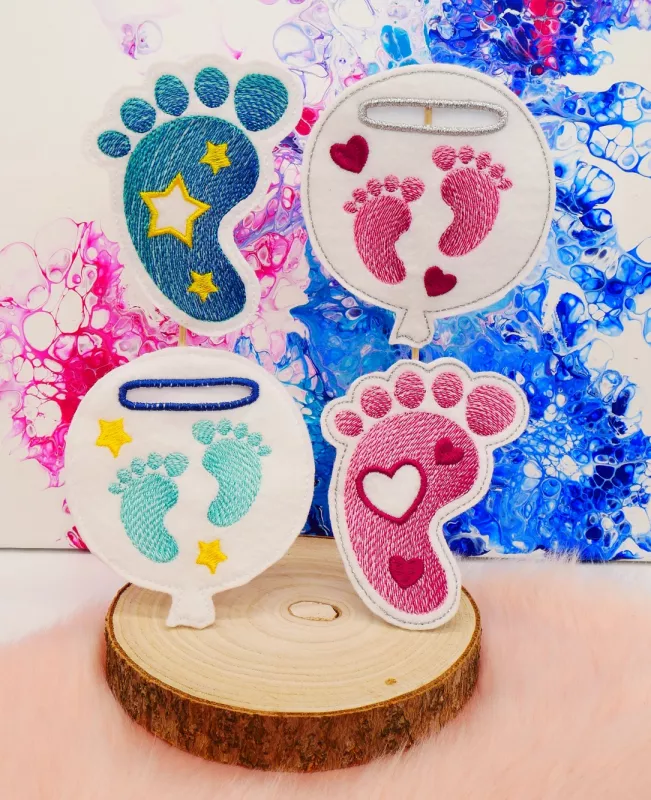 Stickdatei Set Stabdesigns zur Geburt, Foto mit 4 Dekostab-Designs, präsentiert auf Holzscheibe, welche auf orangem Fell liegt. 2 mal Luftballon mit Einstecköse, bestickt mit je einem Paar Füße pink bzw. türkis, umgeben von Herzen bzw. Sternen. 2 mal Fuß