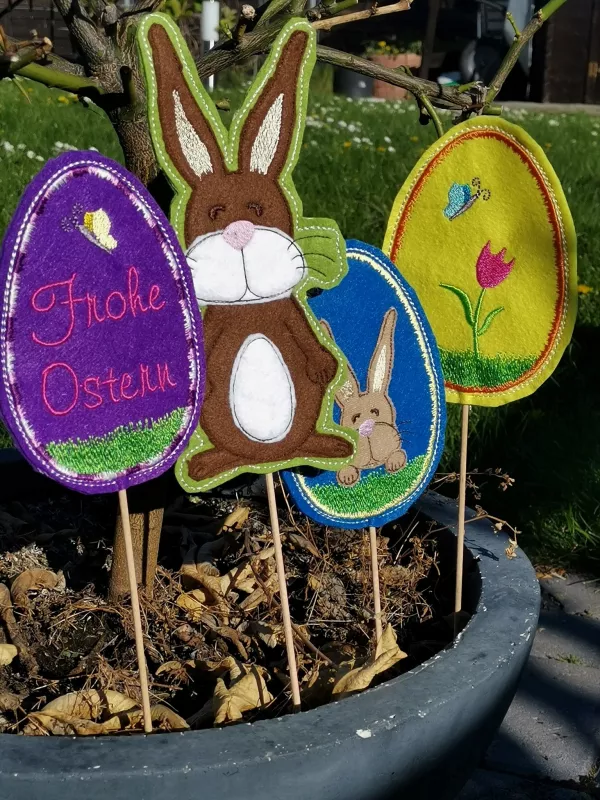 Stickdatei Set ITH-Stecker in Hasen- und Eiform, Foto mit 4 bunten Designstäben in einem Pflanzentopf. 3 mal Osterei (Hasenkopf mit Pfoten auf Gras; Gras mit Tulpe und Schmetterling; Gras mit Schmetterling und Schriftzug "Frohe Ostern"), 1 mal kompletter 