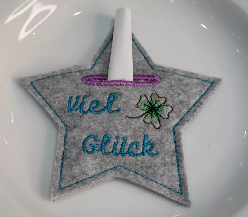 Stickdatei Set Happy new Year Tischdeko (div. Rahmengrößen) , Foto mit Tasche in Sternform auf hellem Teller, in der Einstecköffnung ein gerolltes Papier. Grauer Filz, bestickt mit Schriftzug "Viel Glück" und Glücksklee.