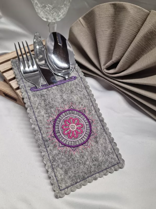 Stickdatei Set Mandala-Bestecktaschen + Einzelmotive, Foto mit Tasche, befüllt mit Gabel/Messer/Löffel, drapiert auf Tischdecke mit anteilig im Hintergrund sichtbarem Glas, bzw. Serviette. Mittelgrau melierter Filz, der am Rand gebuchtet ist und im obere