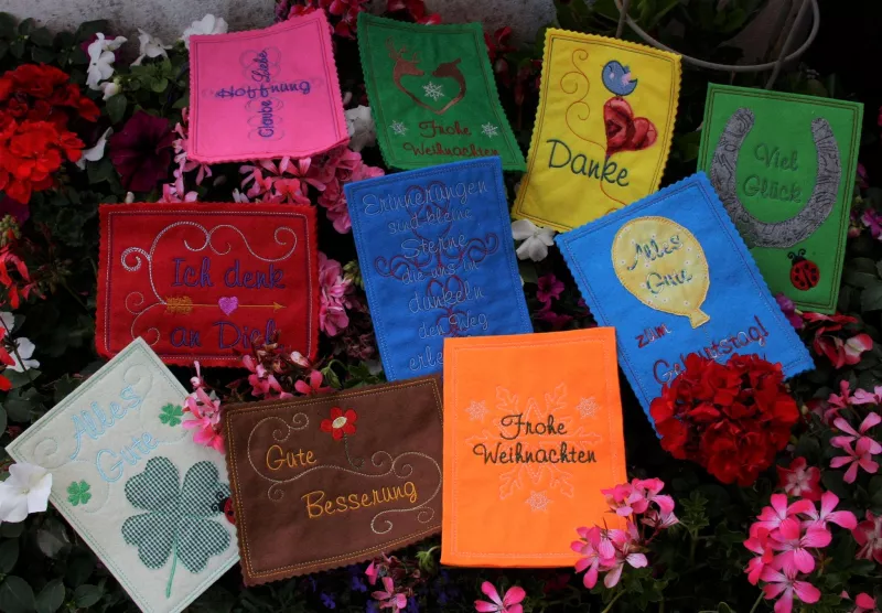 Stickdatei Set ITH Glückwunschkarten inkl. Abwandlung zur Geschenktasche (div. Rahmengr.). Foto von 10 Karten (Hochkant- und Querformat) aus Filz diverser Farben, auf Blumenuntergrund liegend. Bestickungen mit Schriftzügen für diverse Anlässe und jeweils