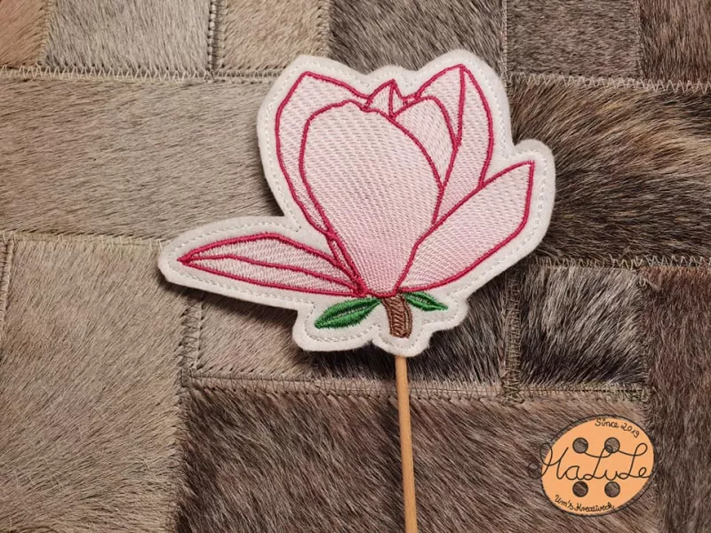 Freebie Stickdatei ITH Stabdatei Magnolie, Foto einer Ausfertigung am Stab, die auf Pelzuntergrund liegt. Untergrundstoff heller Filz in Blütensilhouette. Blütenkonturen rot, Innenfüllung zarte rosa Bestickung, Stilansatz in braun, 2 kleine Blättchen in g