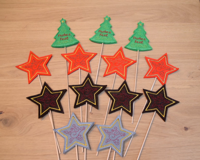 Stickdatei Set ITH Stabdateien mit Engel, Stern und Christbaum, Foto mit 13 Designstäben, auf Holztisch liegend. 10 mal Sterne, unterschiedliche Farben, Spiralverzierung; 3 mal Tannenbaum, Schriftzug "Frohes Fest".