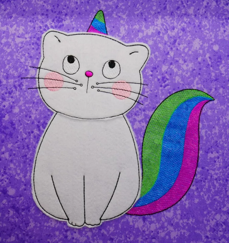 Stickdatei Set KittyCorn Doodle-Applikation inkl. ITH Anhänger, Foto mit Einhorn-Katze, gestickt auf lila meliertem Untergrund. Katze aus hellem Filz, Schwanz und Horn rot-blau-grün gestickt.
