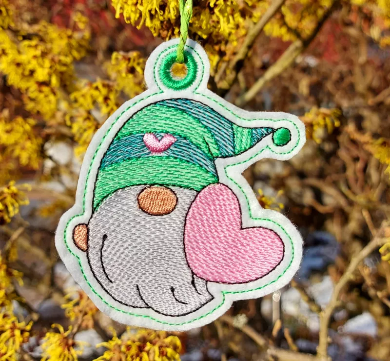 Freebie Stickdatei ITH Anhänger Lovegnome (10x10 Rahmen), Foto zeigt an einer Kordel einen garngeösten Anhänger. Motiv ist ein Gnom mit Mütze in Grüntönen mit rosa Herz und rosa Herz in der Hand.