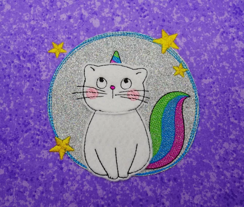 Stickdatei Set KittyCorn Doodle-Applikation inkl. ITH Anhänger, Foto mit in Kreis sitzender Einhorn-Katze, gestickt auf lila gemustertem Untergrund. Einhorn und Schwanz bunt, gelbe Sterne am umgebenden Kreis.