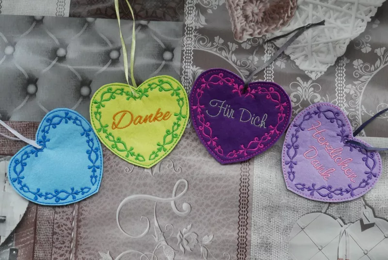 Freebie Danke Herzchen mit ITH Geschenkanhängern (10x10 und 13x18 Rahmen), Foto mit vier Anhängern in Herzform am Bändchen. Untergrund mittelblau mit dunkelblauer schnörkeliger Innenrandbestickung, gelb mit grüner Schnörkelrandbestickung, auberginefarben 