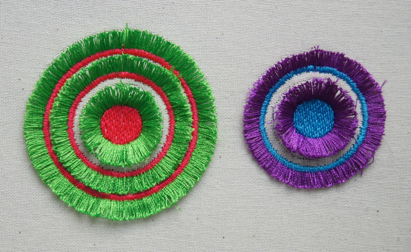 Stickdatei Fransen Set (alle für 10 x 10 cm Rahmen), Foto zeigt auf hellem Stoff zwei kreisrunde Stickmodelle, 1 mal grün-rot, 1 mal lila-blau. Jedes besteht aus einem runden Stickzentrum mit umgebenden Fransen anderer Farbe. Daran anschließend 1, bzw. 2 