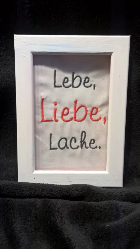 Stickdatei Set Weil ich Dich liebe! (Für 10x10, 13x18 und 18x30), Foto zeigt weißen Bilderrahmen in Hochkantformat. Darin auf hellem Stoff in diversen Farben aufgestickt Schriftzug: "Lebe, Liebe, Lache.".