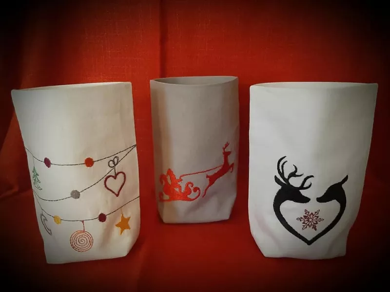 Stickdatei Set 6 winterliche Lichtbeutel (für den 13x18 Rahmen), Foto mit 3 Beuteln aus hellem Stoff. 1 mal Santa-Rentier-Schlitten, 1 mal herzförmig einander zugewandte Rentiere, 1 mal Schnüre mit Weihnachtssymbol-Behang.
