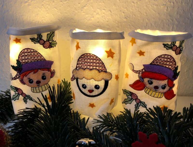 Stickdatei Set ITH Lichtbeutel Christmas Faces, Foto mit 3 hell erleuchteten, am Rand umgeschlagenen hellen Lichtbeuteln mit Motiven. Bestickungen mit Sternen und Stechpalmenzweigen, dabei zusätzlich Schneemannkopf mit Mütze, bzw. Mädchenkopf mit Mütze, b