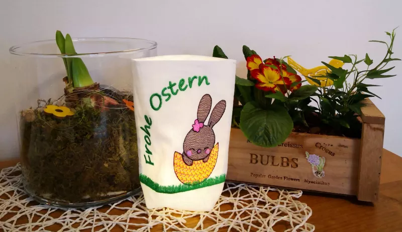 Stickdatei Set Osterhasen ITH Lichtbeutel / Utensilos, Foto zeigt erleuchteten Beutel aus hellem Stoff. Motiv Hase in Eierschale auf Wiese, Schriftzug "Frohe Ostern".