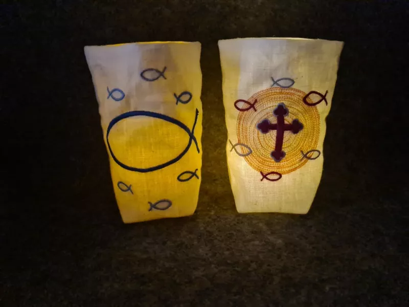 Stickdatei Set ITH Lichtbeutel mit christlichen Motiven, Foto mit hell 2 erleuchteten Ausfertigungen im Dunklen. Beide heller Stoff, eine mit Fischsymbolen groß und klein; eine mit Kreuz in kreisrundem, hellem Schein und kleinen Fischsymbolen.