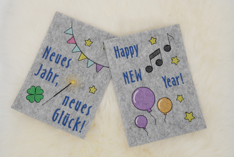 Stickdatei Set Gnom Silvesterparty mit ITH Kartenhüllen & Einzelmotive, Foto zeigt 2 Hochkant-Karten aus hellgrauem Filz. Eine mit Sternen, Girlande, Glücksklee, Wunderkerze, Aufschrift "Neues Jahr, neues Glück!"; eine mit Luftballons, Musiknoten, Sternen